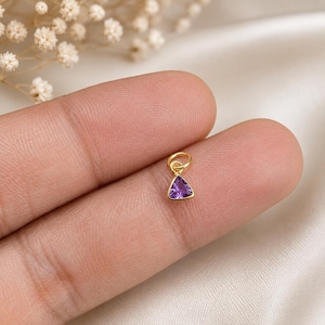 Puede incluir: Un pequeño dije de oro con una piedra preciosa púrpura de forma triangular engastada en un marco de oro pulido, con una argolla circular unida en la parte superior. El dije se muestra sobre un fondo neutro, resaltando el tono violeta intenso.