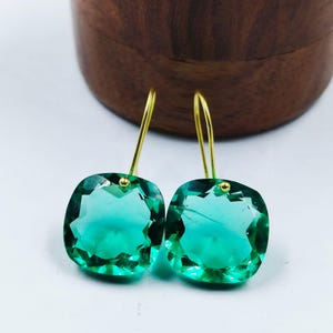 Puede incluir: Un par de pendientes de piedras preciosas verde esmeralda con ganchos dorados. Los pendientes presentan piedras talladas cuadradas y facetadas que reflejan la luz. El fondo incluye un cuenco de madera.