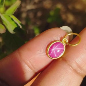Può includere: Un pendente in oro con una gemma rosa ovale con un motivo a stella a sei raggi. La gemma è incastonata in una lunetta d'oro e ha un anello d'oro per una catena. Il pendente è un gioiello.
