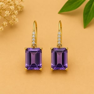 May include: A pair of gold drop earrings featuring rectangular, deep purple gemstones. The earrings have a small row of clear stones on the top part of the hook. The gemstones are set in gold-coloured metal with small gold-coloured accents at each corner.