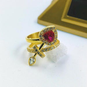 Może przedstawiać: Złoty pierścionek z czerwonym kamieniem w kształcie serca otoczonym małymi, przezroczystymi kamieniami. Pierścionek ma podwójną obrączkę z krzyżem i przezroczystym kamieniem w kształcie łzy.