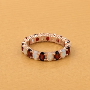 May include: Rose gold ring featuring alternating oval-cut gemstones. The ring showcases a pattern of white opals and deep red garnets. The ring is set against a light brown background.