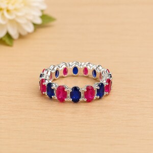 Puede incluir: Un anillo plateado con un patrón alterno de piedras preciosas rosas vibrantes y azul oscuro de corte ovalado. El anillo está sobre una superficie de madera clara, con una flor blanca en el fondo. Las piedras están espaciadas uniformemente.