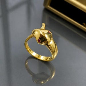 Könnte beinhalten: Ein goldfarbener Ring in Schlangenform mit roten Edelsteinen als Augen. Der Ring hat eine polierte Oberfläche, die das Licht reflektiert. Der Ring ist vor einem dunklen Hintergrund abgebildet, wodurch er gut zur Geltung kommt.