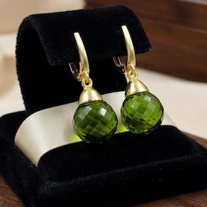 May include: A pair of gold-colored earrings with faceted, olive-green gemstone drops. The earrings are displayed on a black velvet jewelry box with a cream-colored ribbon.