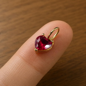 Puede incluir: Un colgante en forma de corazón con una vibrante gema roja engastada en un marco de metal dorado. El colgante tiene un lazo en la parte superior para una cadena. La gema está facetada, captando la luz y creando un efecto brillante. Una joya romántica.