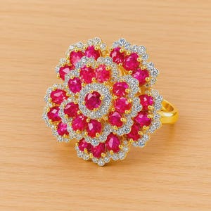 Puede incluir: Un anillo dorado con un diseño grande en forma de flor. Cuenta con numerosas piedras preciosas de color rubí de corte ovalado y piedras preciosas más pequeñas y transparentes. Las piedras están engastadas en un patrón ornamentado, creando una apariencia brillante.