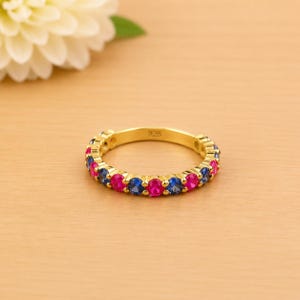 May include: A gold-colored ring with alternating pink and blue gemstones. The ring is set on a light-colored wooden surface. The ring is marked with the number "925". A white flower is in the background.