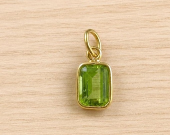 May include: A rectangular, faceted, lime-green peridot gemstone set in a polished yellow gold bezel frame. The pendant features a small, circular gold jump ring attached to the top, allowing it to be suspended from a chain or cord for jewellery.