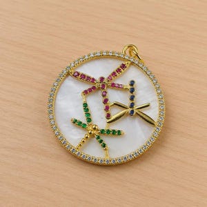 May include: Gold-tone pendant featuring three dragonfly designs with colorful gemstone accents. The dragonflies are adorned with red, green, and blue stones, set against a white background, and surrounded by a ring of clear crystals.