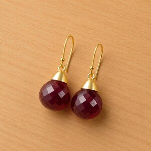 Puede incluir: Un par de pendientes colgantes dorados con colgantes de piedras preciosas rojo oscuro facetadas. Los pendientes tienen un diseño sencillo y elegante, con una tapa dorada que conecta la piedra preciosa con el gancho dorado.