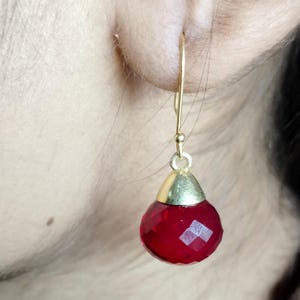 Puede incluir: Un solo pendiente dorado con una gema roja facetada en forma de lágrima. La gema está rematada con metal dorado. El pendiente cuelga de un gancho de alambre dorado.