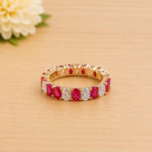 May include: A gold eternity ring with alternating oval-cut red and clear gemstones. The ring is set on a light-coloured wooden surface, with a blurred white and yellow flower in the background.