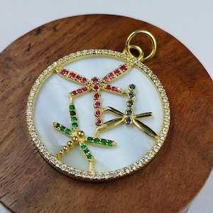 Puede incluir: Colgante dorado con tres diseños de libélulas. Las libélulas están adornadas con piedras preciosas rojas, verdes y azules. El colgante tiene un fondo blanco de nácar y está rodeado por un anillo de piedras preciosas transparentes. Hay un lazo dorado en la parte superior.