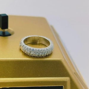 May include: A silver ring with two rows of clear, sparkling stones. The ring is set on a gold-colored surface. The ring's design is simple yet elegant, making it a versatile piece of jewelry.