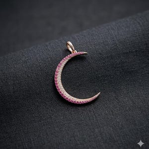Op de afbeelding: Een maan-hanger in de vorm van een halve maan met een roségouden afwerking. De maan is bezet met roze en witte edelstenen. De hanger wordt gepresenteerd op een donkergrijze stof.