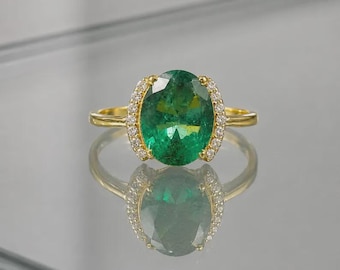 May include: A gold ring featuring a large, oval-cut emerald gemstone. The emerald is surrounded by a halo of small, clear gemstones. The ring's band is a simple, polished gold.