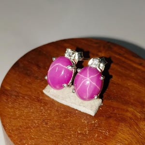 Op de afbeelding: Een paar oorstekers met grote, ovale, magenta edelstenen met een stervormig patroon. De oorbellen zijn gezet in zilver metaal met kleine accentstenen. De oorbellen worden gepresenteerd op een kleine, witte, rechthoekige basis.