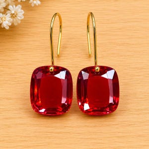 May include: A pair of red, square-cut gemstone earrings with gold-colored hooks. The earrings are displayed on a light brown wooden surface, with white flowers in the background.