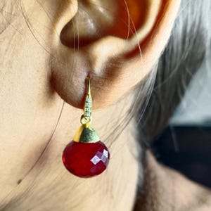 Puede incluir: Un pendiente de color dorado con una piedra roja facetada en forma de lágrima. El pendiente tiene una pequeña sección decorativa con piedras transparentes cerca del lóbulo de la oreja. El pendiente cuelga de una oreja.