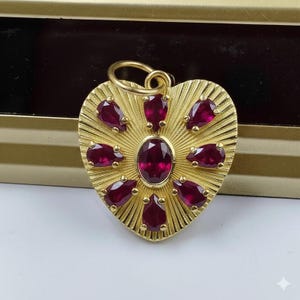 May include: A gold heart pendant featuring a central oval ruby and surrounding pear-shaped rubies. The pendant has a textured, sunburst design and a loop for a chain. The rubies are a deep red colour.