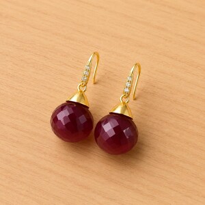 Puede incluir: Un par de pendientes colgantes con esferas de piedras preciosas facetadas de color rojo intenso. Los pendientes tienen ganchos dorados con pequeños acentos brillantes y tapas doradas. Se exhiben sobre una superficie de madera marrón claro.