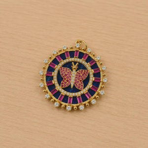 May include: A gold-colored pendant with a butterfly design. The pendant features a central butterfly shape with pink accents, surrounded by a blue and pink circular pattern. The outer edge is adorned with small, clear gemstones.