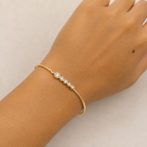 May include: A gold bangle bracelet featuring five clear, round gemstones. The gemstones are set in a line along the top of the bracelet. The bracelet is on a person's arm.