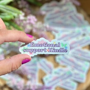 May include: A blue and white sticker with the text "Emotional support Kindle" in a retro font. The sticker has a floral design with butterflies and stars.