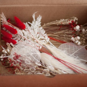 May include: A cardboard box filled with dried flowers and foliage. The box contains white fern fronds, white dried leaves, red dried flowers, and white seed pods.