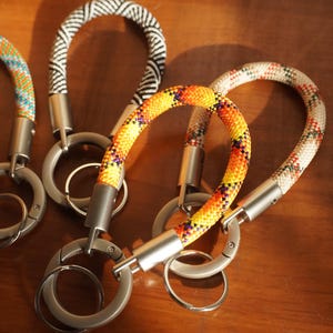 May include: A collection of four circular keychains made from braided climbing rope in various patterns, including black and white chevron, bright orange and yellow, and multicoloured designs, each attached to a matte silver metal ring and clasp.