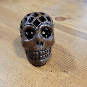 May include: A detailed, brown skull figurine with a lattice design on top. The skull has large eye sockets and a detailed jawline. The figurine is made of a dark brown material and sits on a wooden surface.