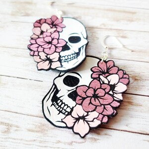 May include: A pair of dangle earrings featuring a white skull with black details and pink flowers. The skull is partially covered by the flowers.