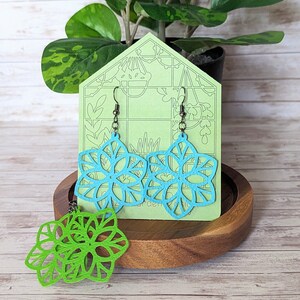 May include: A pair of blue and green laser cut earrings in the shape of a flower. The earrings are displayed on a green card with a greenhouse design. The card is sitting on a wooden coaster.