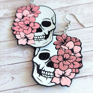 May include: A pair of dangle earrings featuring a white skull with pink flowers covering one eye. The earrings are made of leather and have silver hooks.