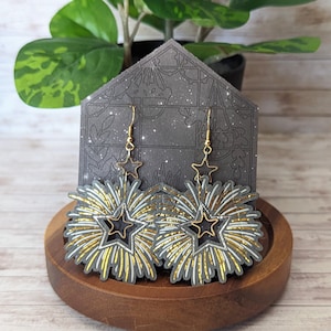 May include: A pair of gold and black starburst earrings with a glitter finish. The earrings are hanging from a gold hook.