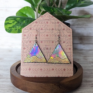 May include: A pair of iridescent triangle earrings with a gold glitter wave design. The earrings are displayed on a brown card with the text "Rose Garden Design".