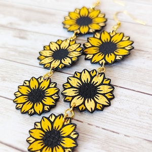 May include: A pair of dangle earrings featuring three gold-toned sunflower charms with black centers. The sunflowers are layered and connected by gold-toned chains.