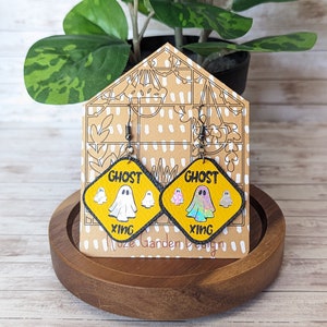 May include: A pair of yellow and black ghost crossing sign earrings with a holographic ghost design. The earrings are on a brown wooden display stand. "Ghost Xing" is written on the signs.