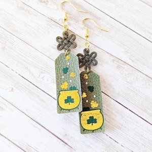 May include: A pair of green and gold dangle earrings with a pot of gold charm and a Celtic knot design. The earrings are made of leather and have gold-tone hooks.