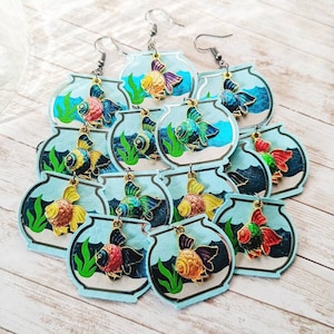 May include: A set of 12 colorful fishbowl earrings. Each earring features a different colored fish with a gold trim. The fishbowls are blue with green seaweed and white sand.