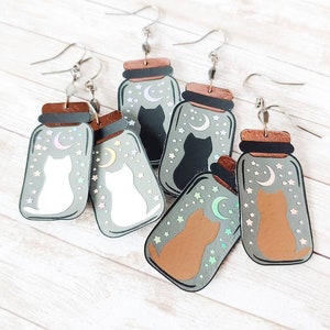 May include: Four pairs of cat-themed earrings. Each earring features a glass jar with a cat inside, a crescent moon, and stars. The cats are white, black, brown, and a light pink color. The jars are a light green color with a brown lid.