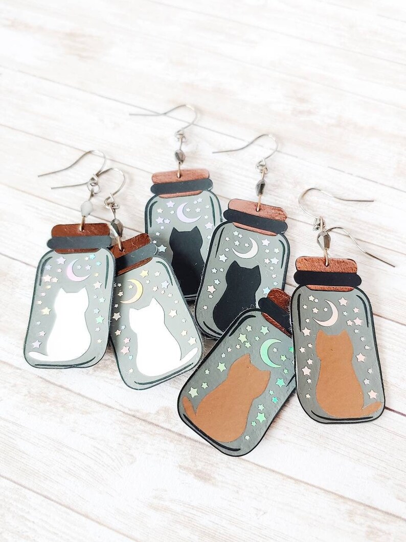 May include: Four pairs of cat-themed earrings. Each earring features a glass jar with a cat inside, a crescent moon, and stars. The cats are white, black, brown, and a light pink color. The jars are a light green color with a brown lid.