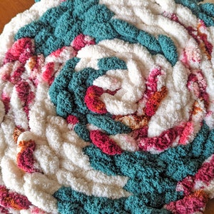 Op de afbeelding: Handgemaakt, cirkelvormig vloerkleed met een spiraalvormig ontwerp. Het vloerkleed heeft een combinatie van wit, teal, roze en oranje dik garen. De textuur is zacht en pluche, waardoor een gezellige uitstraling ontstaat. Geschikt voor interieurdecoratie.