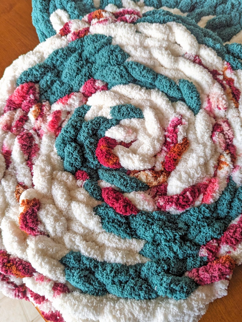 Op de afbeelding: Handgemaakt, cirkelvormig vloerkleed met een spiraalvormig ontwerp. Het vloerkleed heeft een combinatie van wit, teal, roze en oranje dik garen. De textuur is zacht en pluche, waardoor een gezellige uitstraling ontstaat. Geschikt voor interieurdecoratie.
