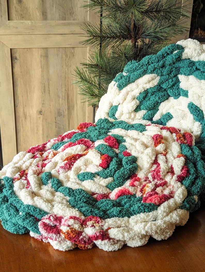 Op de afbeelding: Twee ronde kussens van dikke breisels in een spiraalpatroon. De kussens hebben een combinatie van wit, blauwgroen, roze en oranje garen. De kussens liggen op een houten oppervlak met een kunsttak van een dennenboom op de achtergrond.