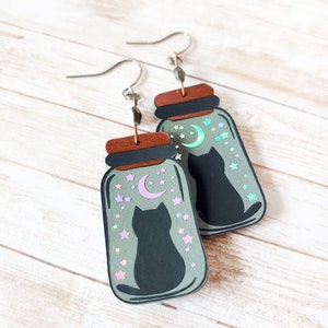 May include: A pair of dangle earrings featuring a black cat silhouette inside a glass jar with a crescent moon and stars. The jar has a holographic finish with iridescent colors.