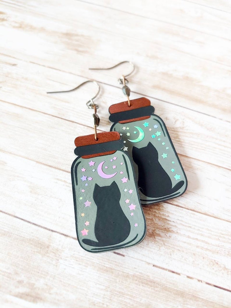May include: A pair of dangle earrings featuring a black cat silhouette inside a glass jar with a crescent moon and stars. The jar has a holographic finish with iridescent colors.