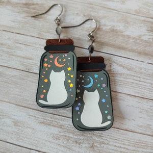 May include: A pair of cat-themed earrings featuring a white cat silhouette inside a glass jar with a crescent moon and stars. The jar is painted in a dark gray color with iridescent rainbow accents.