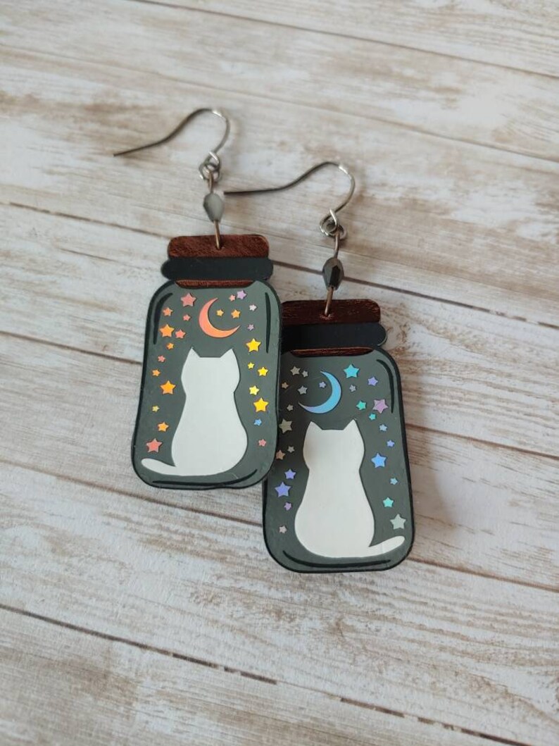 May include: A pair of cat-themed earrings featuring a white cat silhouette inside a glass jar with a crescent moon and stars. The jar is painted in a dark gray color with iridescent rainbow accents.